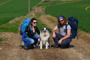 Couple de randonneurs avec sacs à dos posant avec leur chien sur un chemin de campagne entouré de champs verdoyants, lors d’une pause en pleine nature.