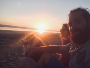 Un couple assis sur la plage au coucher du soleil avec leur chien, moment chaleureux et complice en bord de mer.